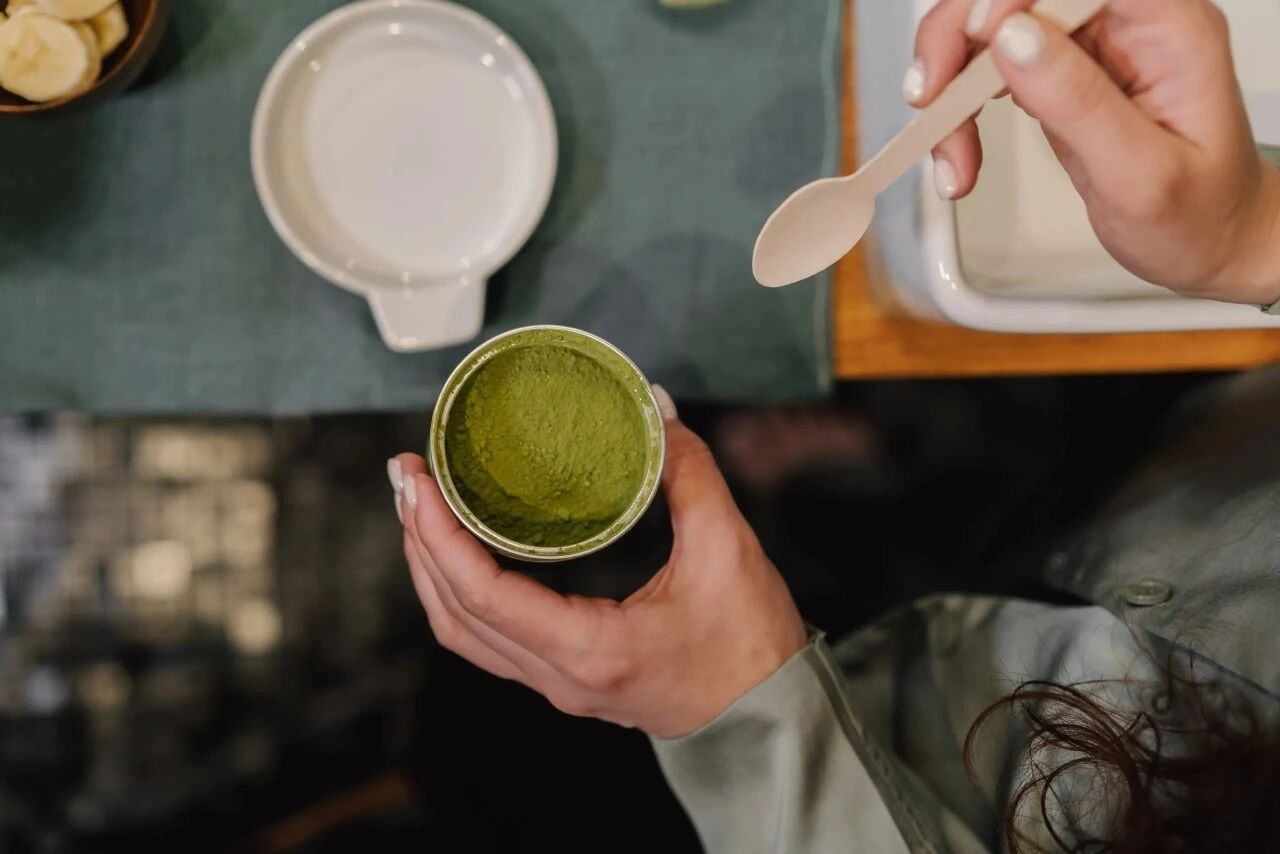 Hand holding a tin of bright green matcha powder with a spoon, ready for food and beverage applications.