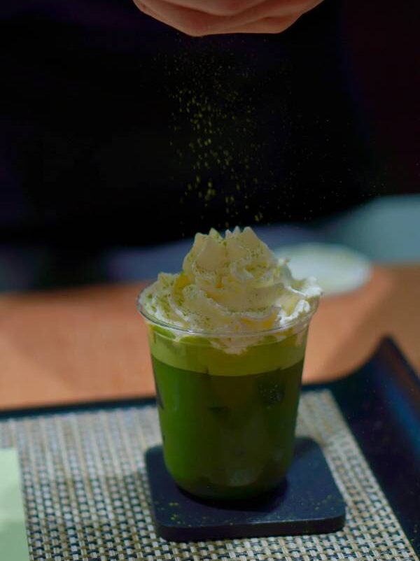 Hand dusting matcha powder over an iced jasmine matcha cream cap with whipped cream topping.