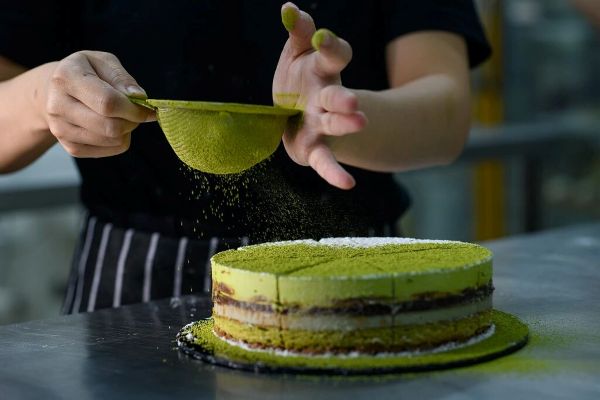 Chef dusting matcha powder over a layered cake, showing how matcha is used in bakery and dessert products.